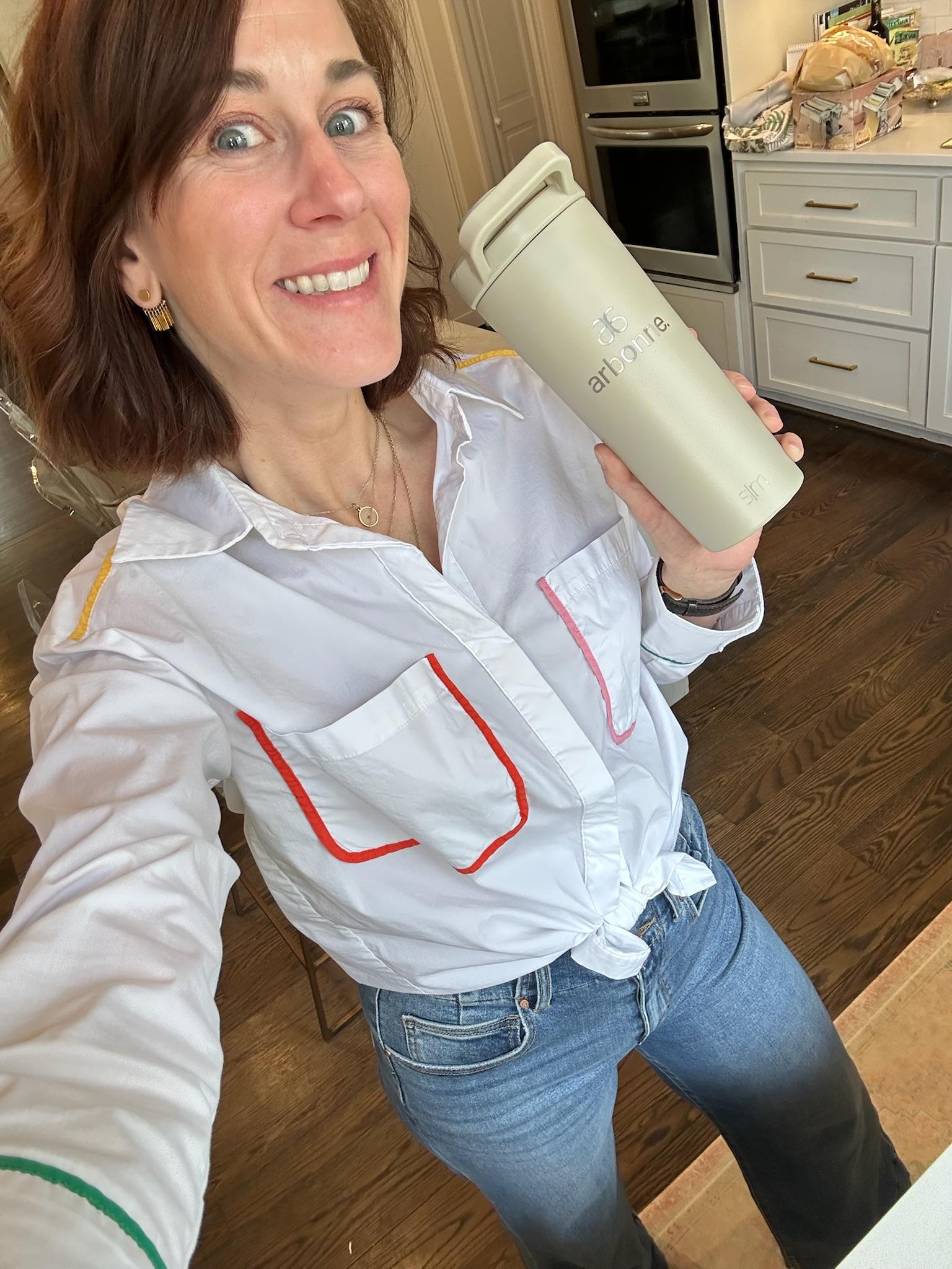 Louise in a white shirt with coloured topstitching, holding a sage green tumbler in her kitchen